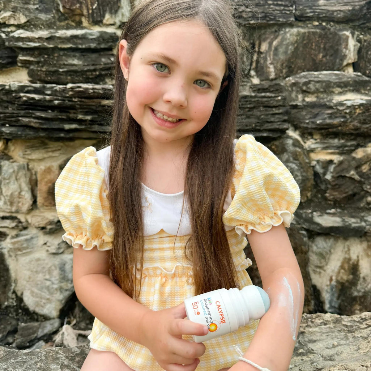 Refect Beauty Calypso Kids SPF50 roll-on sunscreen being applied by a young girl in a yellow gingham dress