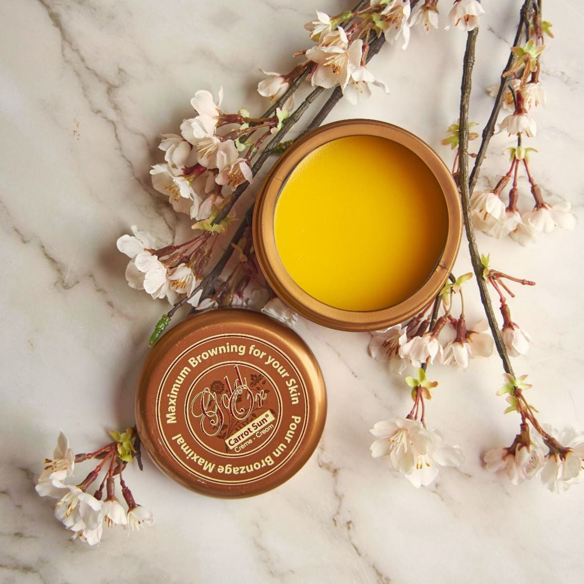 Refect Beauty Carrot Sun Gold tanning cream in an open bronze tin with white cherry blossom branches on marble