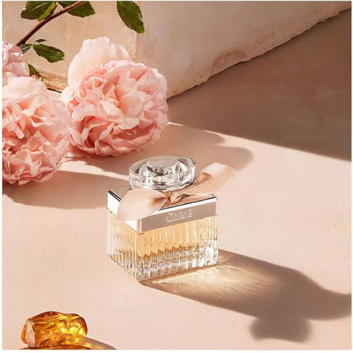 Refect Beauty Chloé Eau de Parfum bottle with a beige ribbon, surrounded by pink peonies on a warm-toned surface.