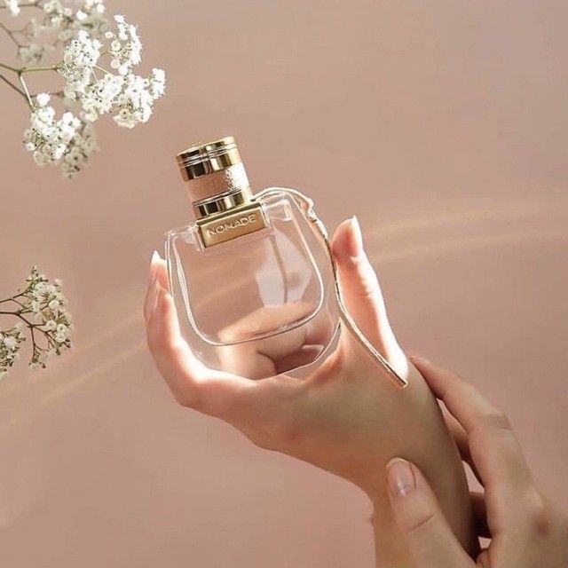 Refect Beauty Chloé Nomade Eau de Parfum glass bottle held in a hand against a soft pink background with baby's breath
