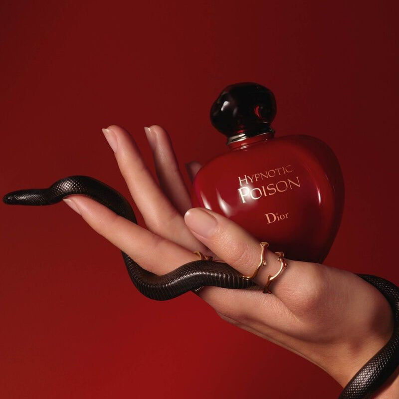 Refect Beauty Dior Hypnotic Poison perfume bottle held in a hand with a black snake against a deep red background.