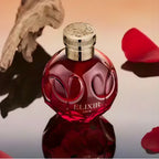 Refect Beauty Elie Saab Elixir Love perfume bottle in deep red glass with gold cap, surrounded by rose petals.