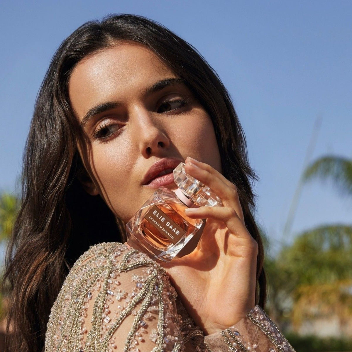 Refect Beauty model holding Elie Saab Le Parfum Essentiel bottle, wearing an elegant embellished dress outdoors.