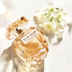 Refect Beauty Elie Saab Le Parfum Lumière perfume bottle with golden liquid and white jasmine flowers in sunlight.