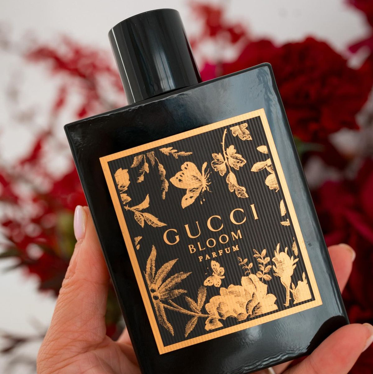 Refect Beauty Gucci Bloom Parfum bottle with black glass and gold floral label held in a hand against red flowers