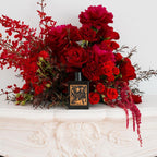 Refect Beauty Gucci Bloom Parfum bottle displayed in front of a lush arrangement of deep red flowers on a mantelpiece.
