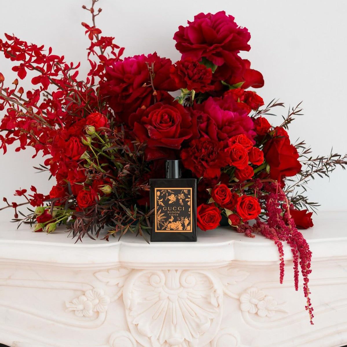 Refect Beauty Gucci Bloom Parfum bottle displayed in front of a lush arrangement of deep red flowers on a mantelpiece.