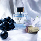 Refect Beauty Guess 1981 Indigo perfume bottle with fresh grapes, a wine cork, and a lilac flower on white fabric.