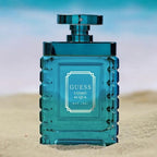 Refect Beauty Guess Uomo Acqua Eau de Toilette in a blue glass bottle on a sandy beach with an ocean background