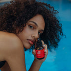 Refect Beauty Hair Addict The Swim Proof Recipe spray held by a woman with curly hair by a swimming pool