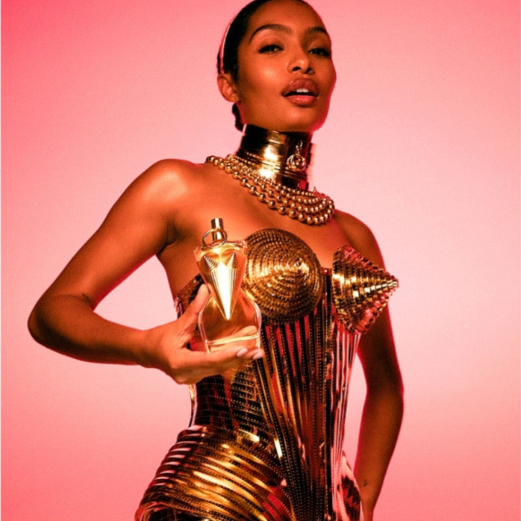 Refect Beauty Jean Paul Gaultier Divine perfume bottle held by a model in a gold metallic corset against a pink background