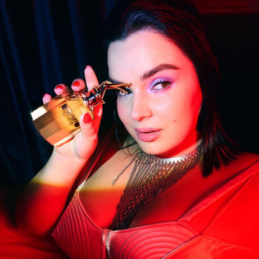 Refect Beauty model holding a gold Jean Paul Gaultier Scandal Absolu perfume bottle in dramatic red lighting