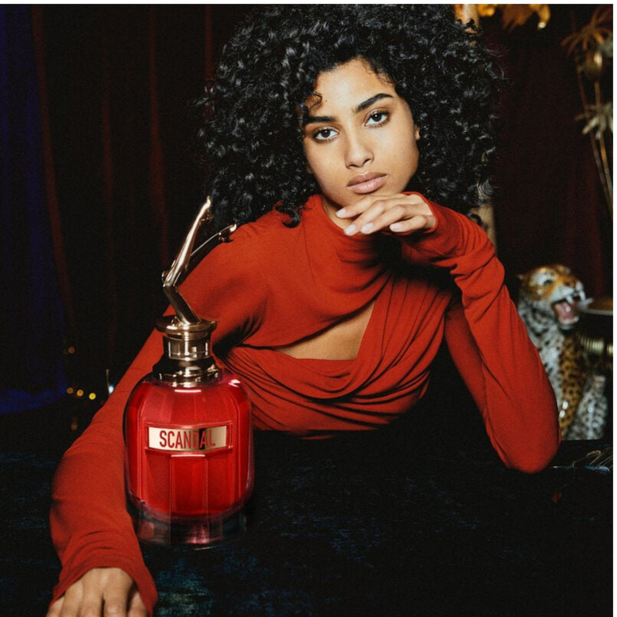 Refect Beauty model with curly hair wearing a red top next to a Jean Paul Gaultier Scandal Le Parfum bottle