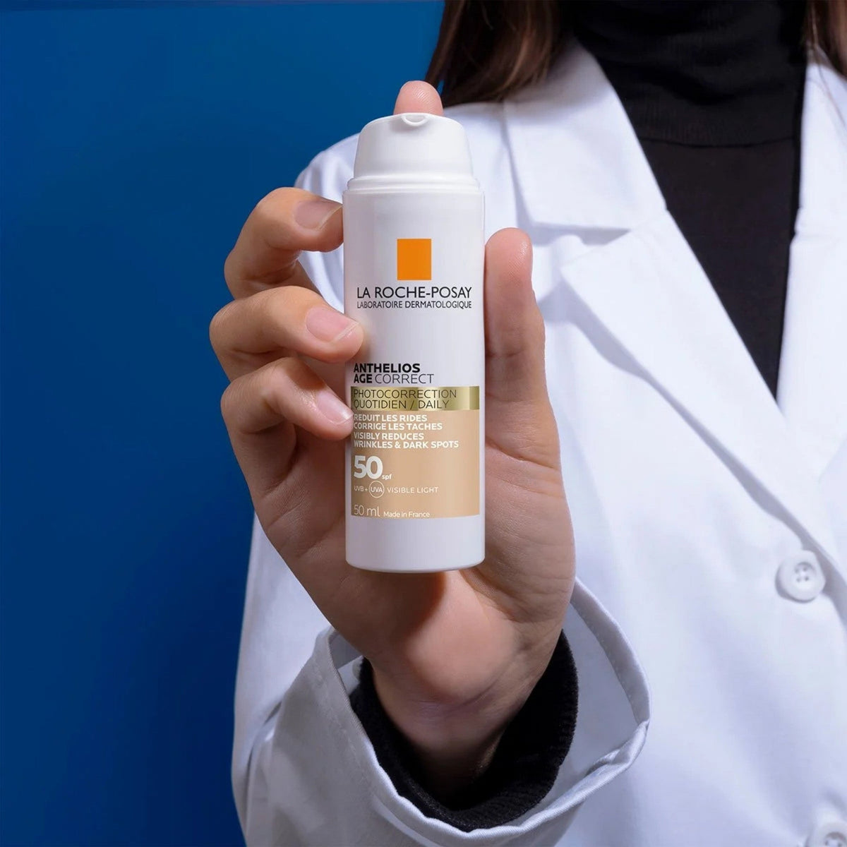 Refect Beauty hand holding a bottle of La Roche-Posay Anthelios Age Correct SPF50 CC cream against a blue background.