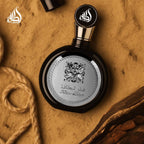 Refect Beauty Lattafa Fakhar Eau de Parfum in a round black bottle with silver ornate detailing on a sandy background.