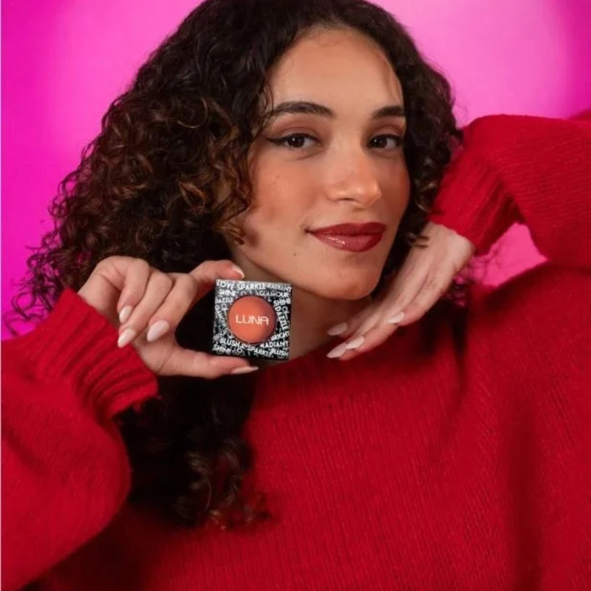 Refect Beauty LUNA Blusher powder compact held by a woman with curly hair wearing a red sweater against a pink backdrop