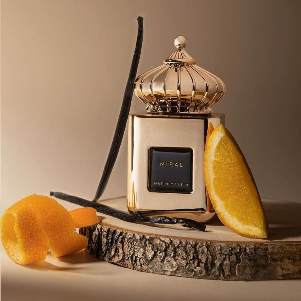 Refect Beauty Matin Martin Miral perfume bottle with gold finish, vanilla beans, and orange slices on wood slice.