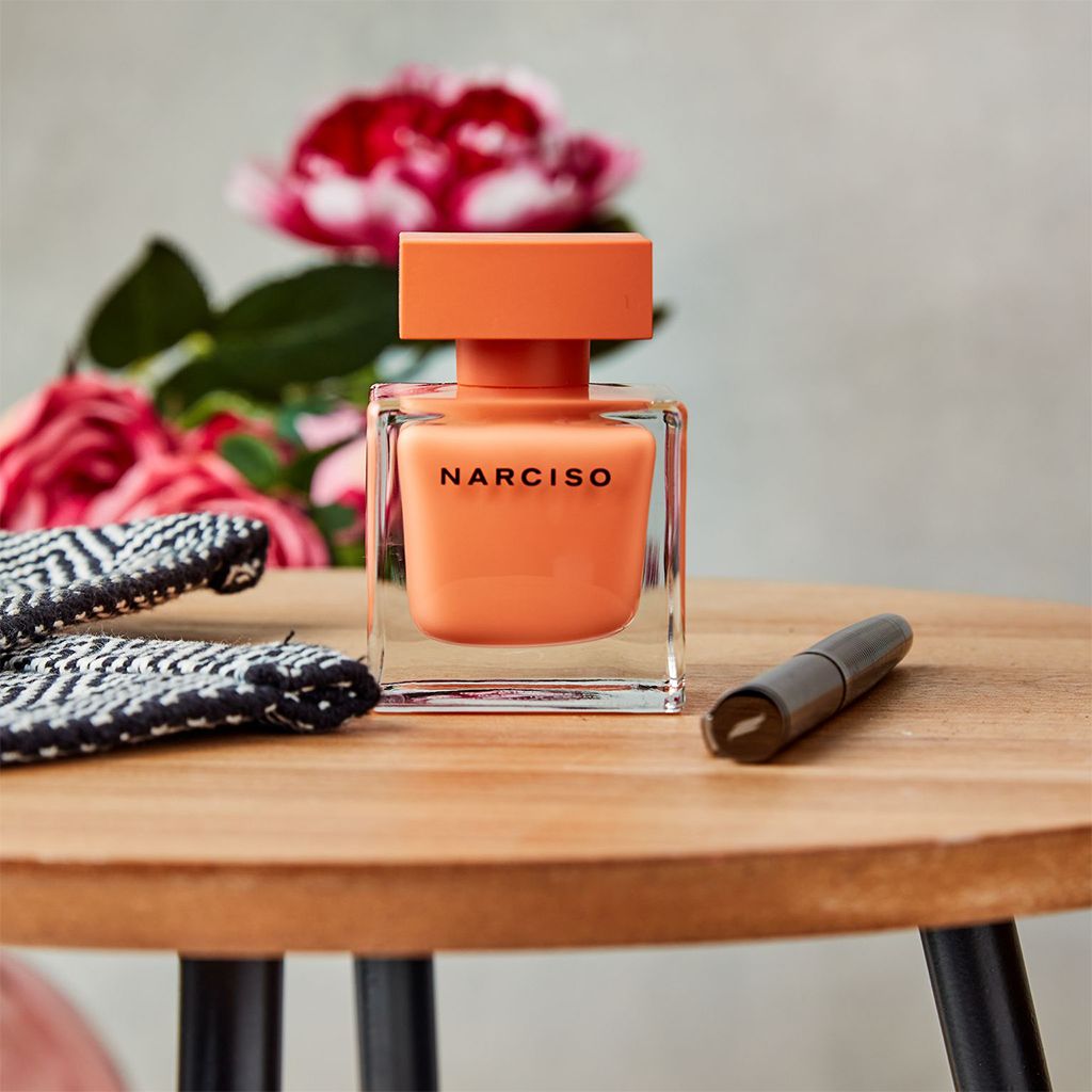 Refect Beauty Narciso Ambree Eau de Parfum in a square peach bottle on a wooden table with flowers in the background