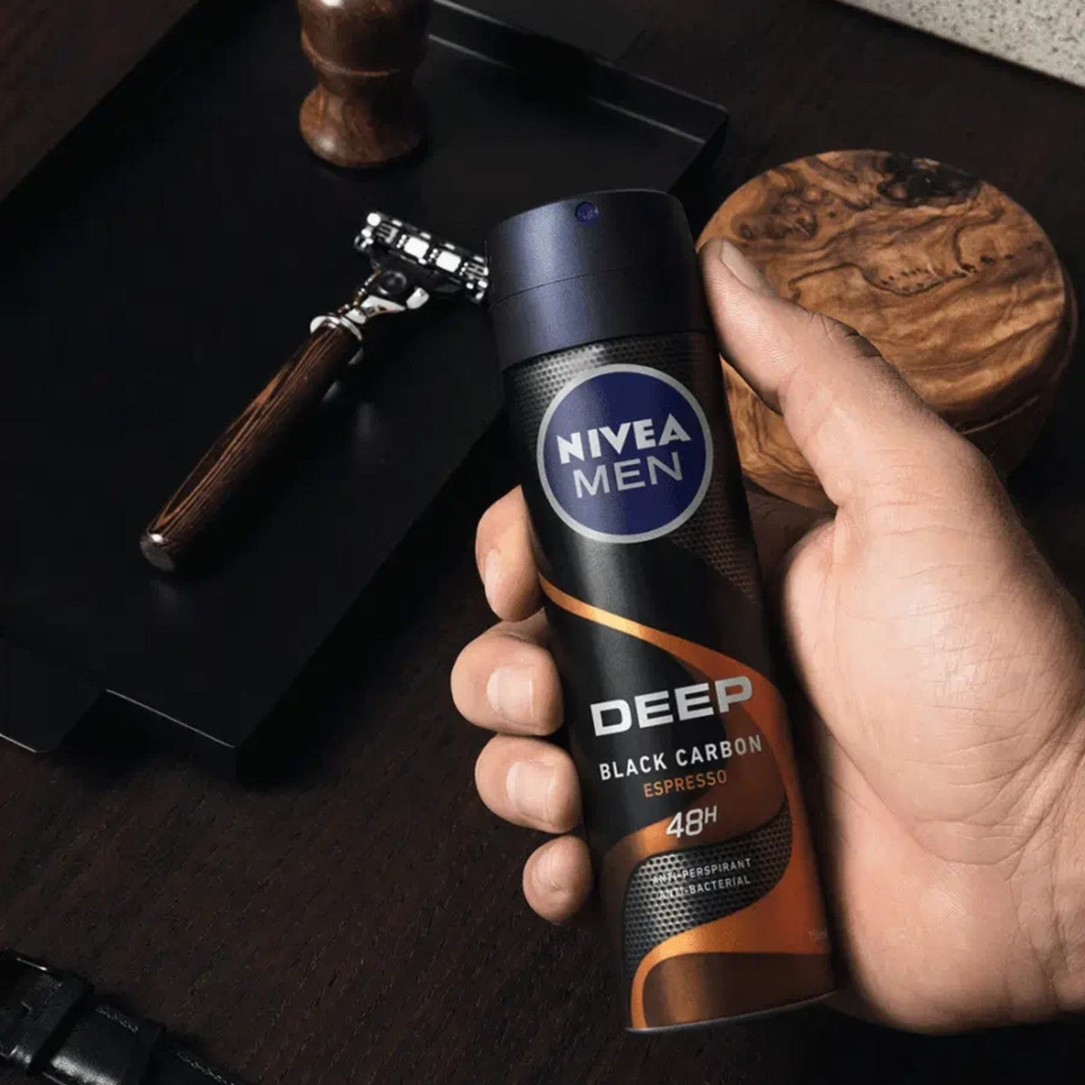 Refect Beauty Nivea Men Deep Espresso body spray held in a hand with grooming accessories on a dark wooden table.