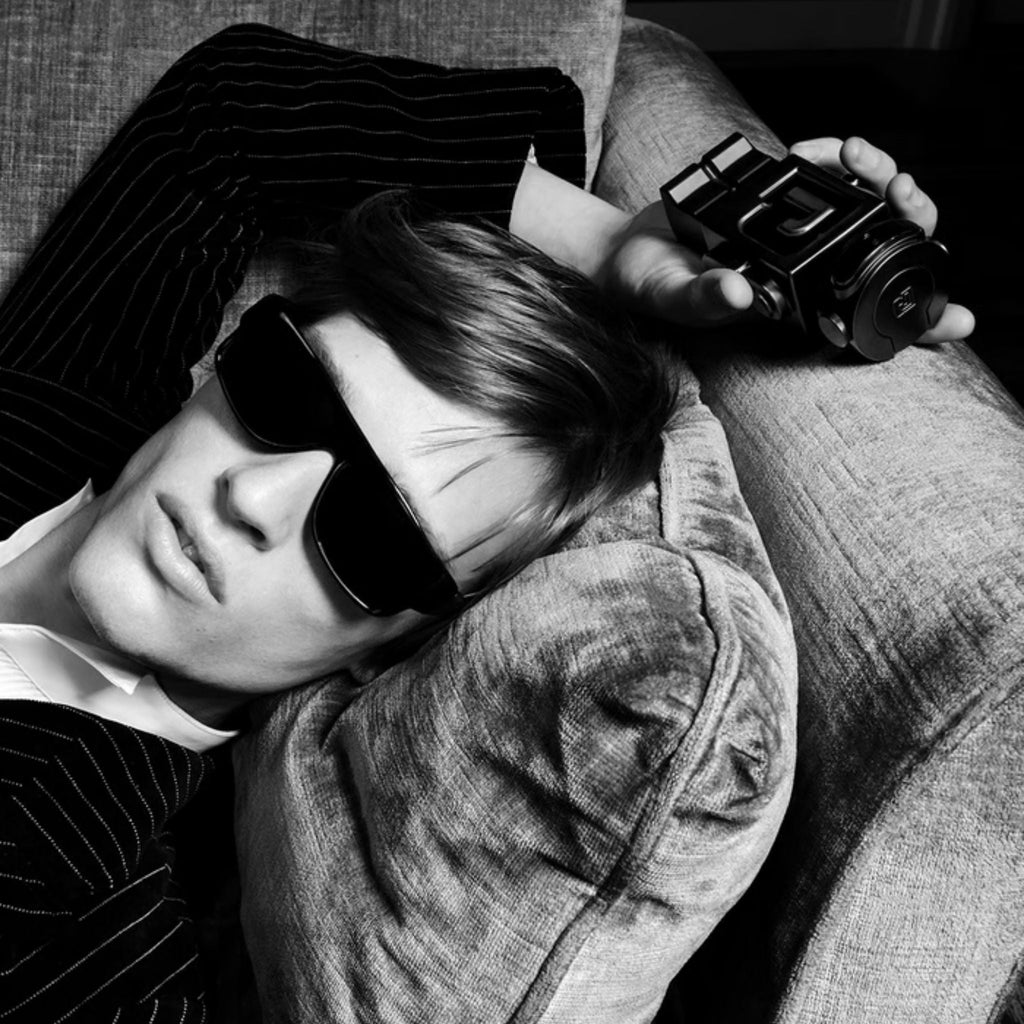 Refect Beauty model wearing sunglasses and a pinstripe suit, holding a Paco Rabanne Phantom Parfum bottle on a sofa.