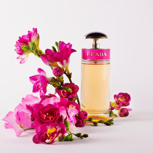 Refect Beauty Prada Candy Eau de Parfum bottle with a pink leather band next to vibrant pink and yellow flowers