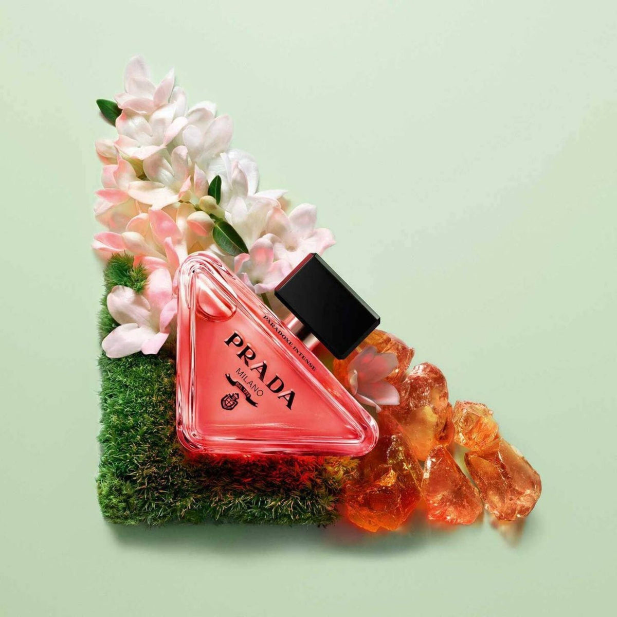 Refect Beauty Prada Paradoxe Intense perfume bottle surrounded by pink flowers, moss, and amber on a green background.
