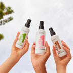 Refect Beauty hands holding three Revlon Uniq One leave-in hair treatment spray bottles against a bright sky