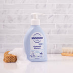 Refect Beauty Sanosan Baby Chamomile Shampoo bottle with pump dispenser, foam, and a wooden brush on a white surface.