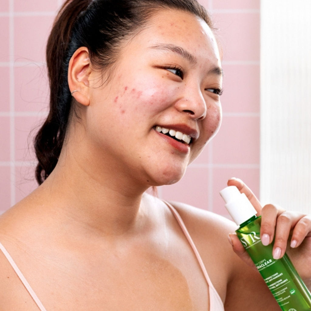 Refect Beauty SVR Sebiaclear body spray held by a woman with acne-prone skin in a pink tiled bathroom